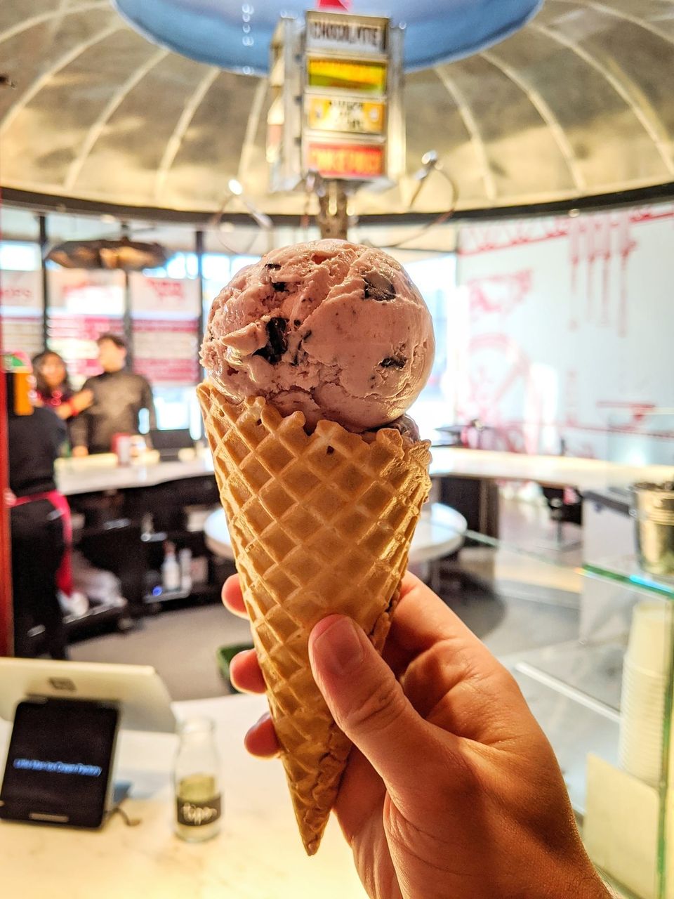 Hand holding a waffle cone with a pink chocolate chip scoop inside an ice cream shop