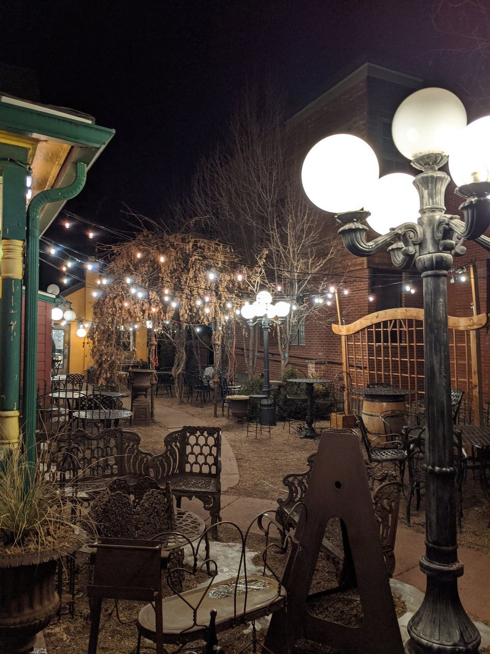 Café patio at night with string lights, globe lamp posts and ornate iron chairs beside a brick building