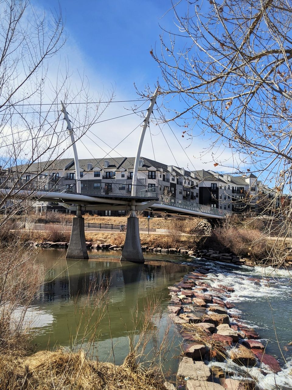 White cable-stayed pedestrian bridge over a river with stone rapids and apartment buildings behind
