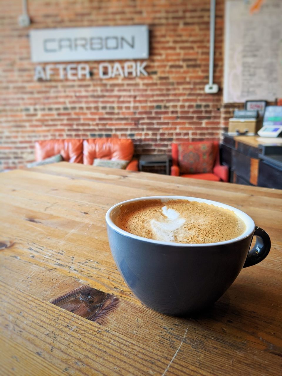 Latte in a gray cup on a wood table with a Carbon After Dark sign on a brick wall behind