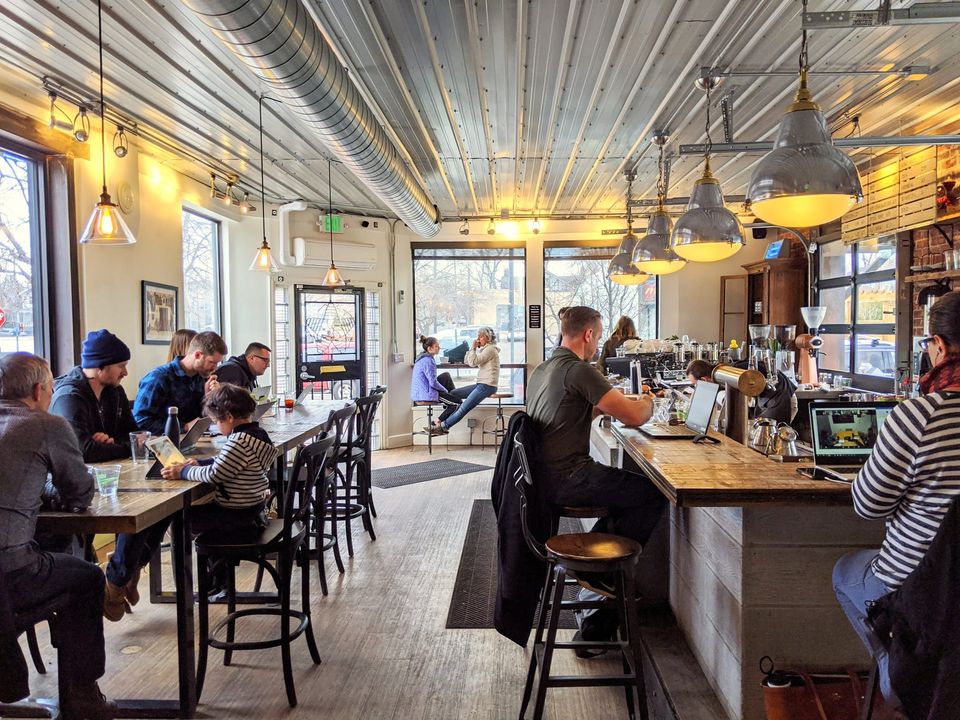 Busy coffee shop with a corrugated metal ceiling, pendant lights and people working at long tables and the bar
