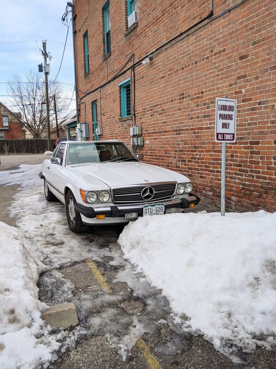 White vintage Mercedes convertible parked in snow beside a brick wall and a Landlord Parking Only sign