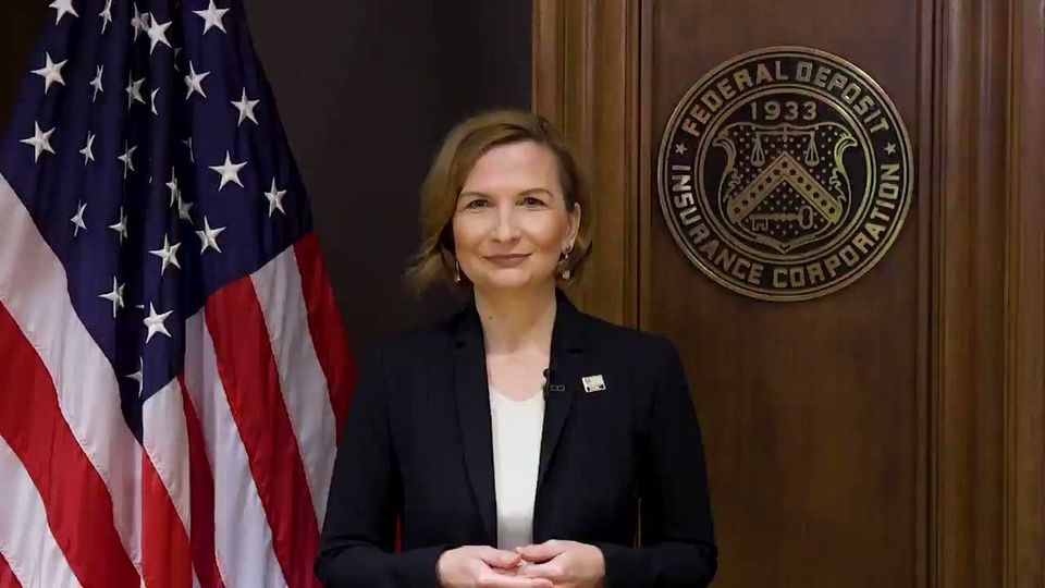 A woman in a dark blazer standing between an American flag and the FDIC seal