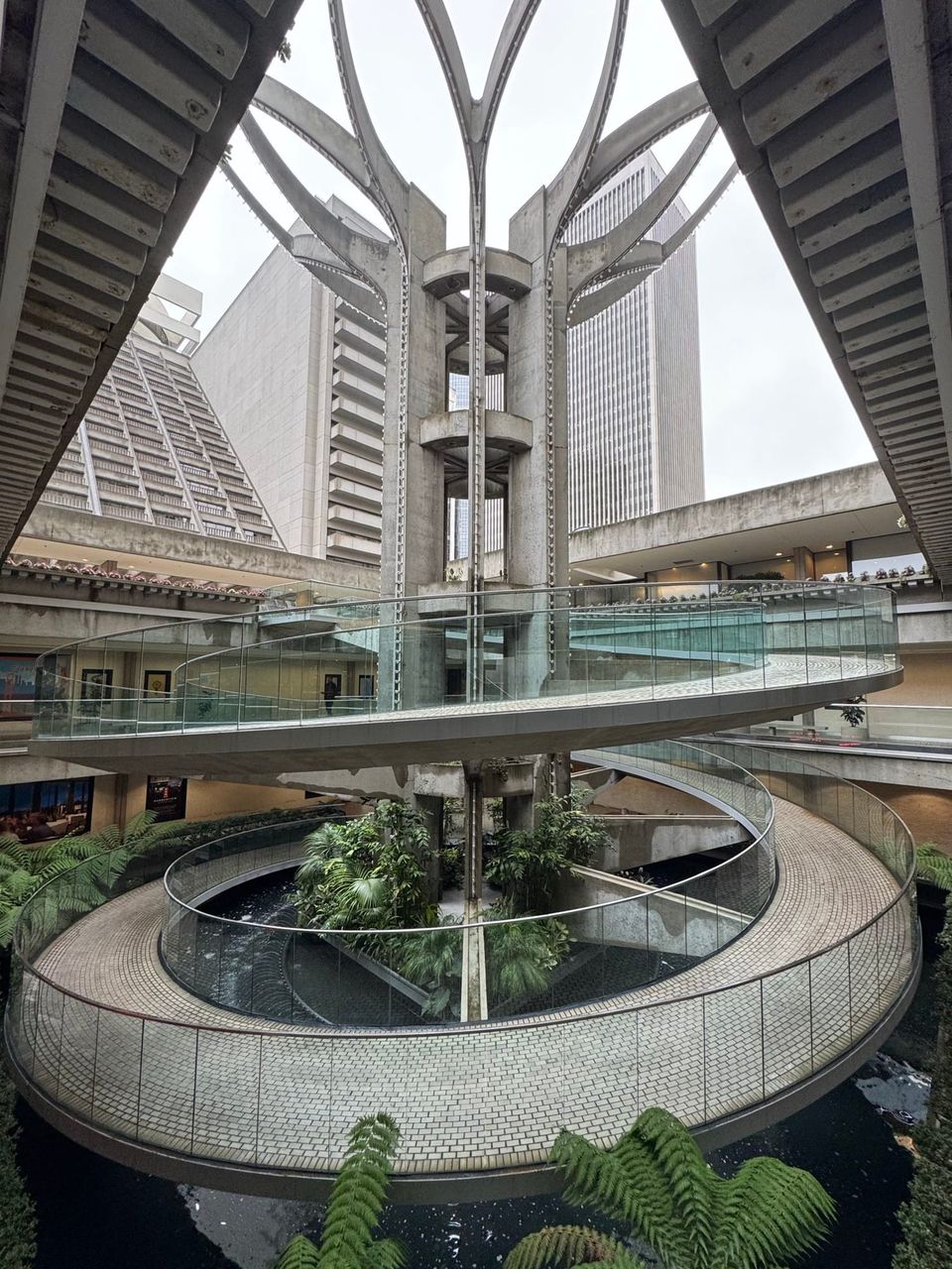 Spiral glass-railed walkway around a concrete tower above a fern-filled atrium surrounded by high-rises