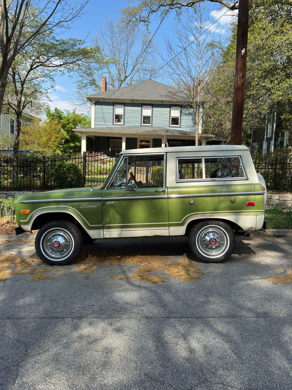 Side profile of a green and white vintage Ford Bronco parked in front of a blue two-story house