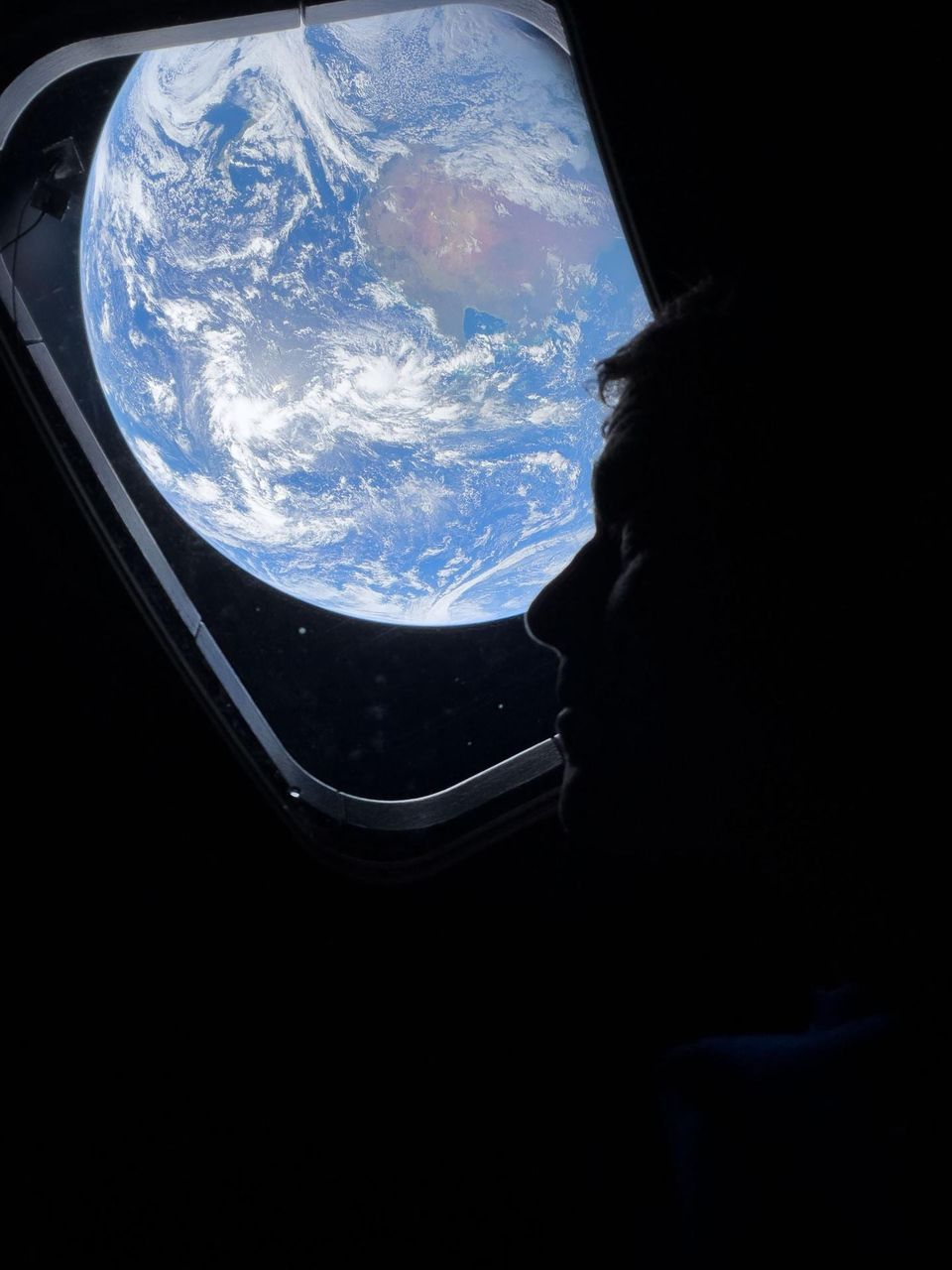 Silhouetted astronaut looking out a spacecraft window at a blue cloud-covered Earth
