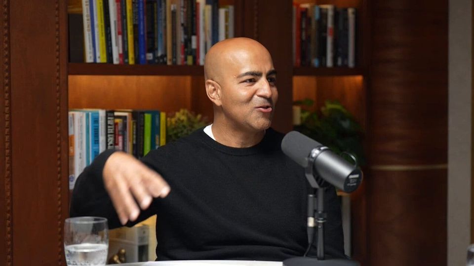 Bald man in a black sweater gesturing while talking into a podcast microphone in front of bookshelves