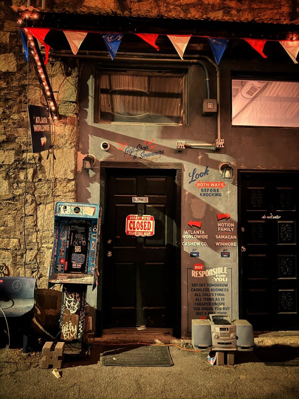 Shop door with a Sorry! We're Closed Please Call Again sign, painted directional signs, a sticker-covered box and boombox