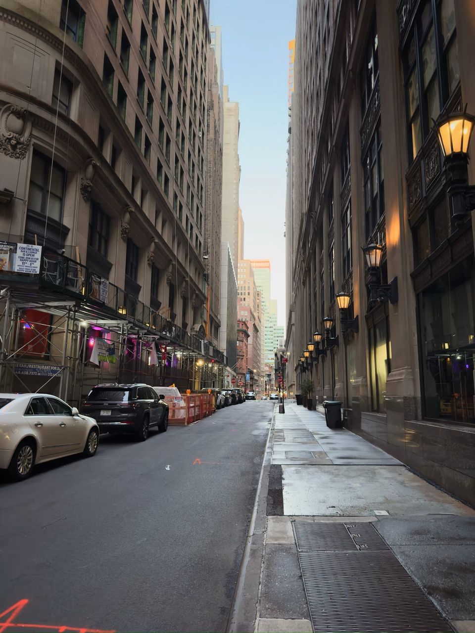 Narrow Financial District street with parked cars, scaffolding and glowing lanterns