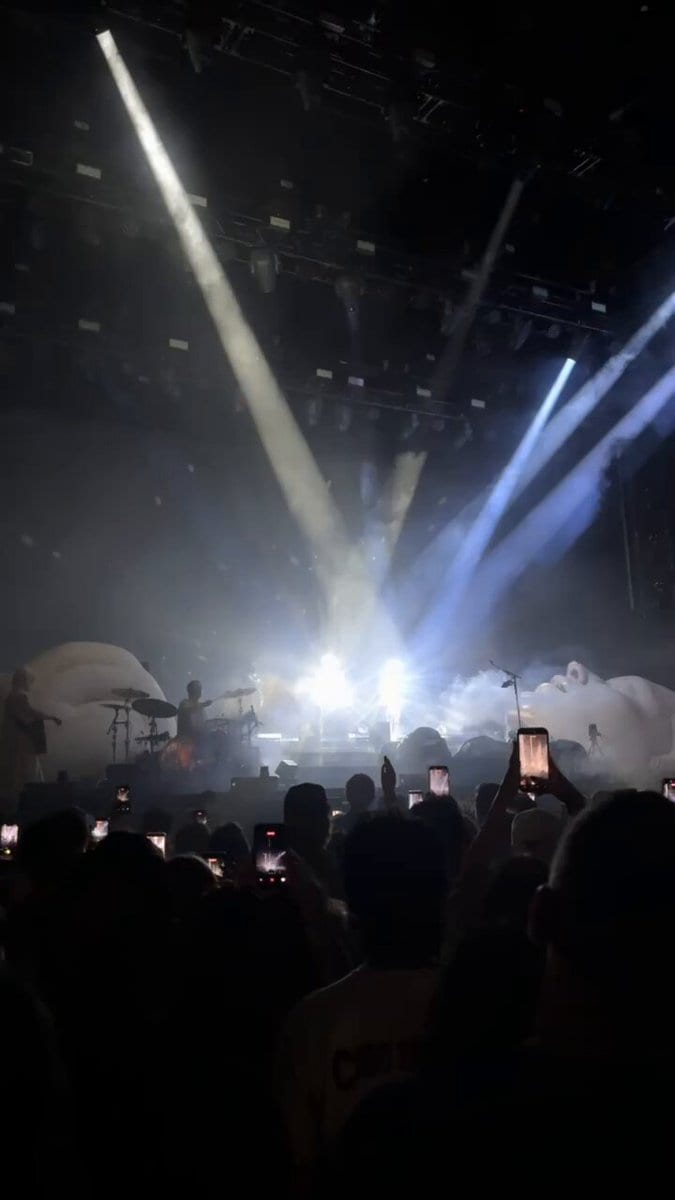 Concert stage with bright spotlights and giant sculpted faces as the crowd holds up phones