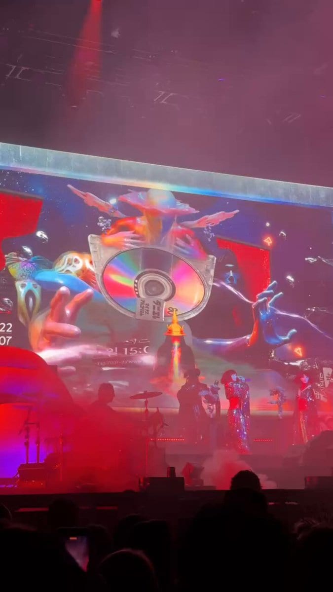 Performers in sparkling costumes before a psychedelic screen showing a giant CD and hands