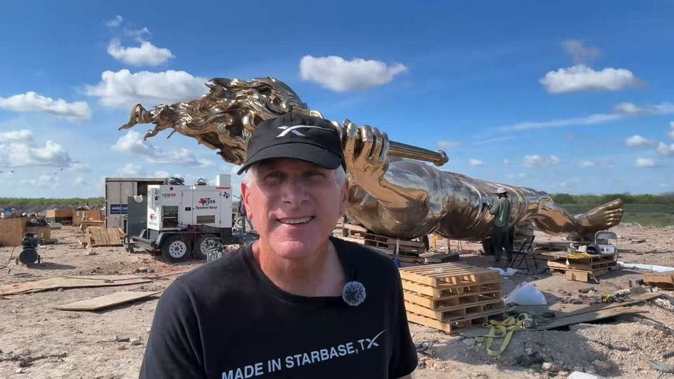 A man in a black cap and “MADE IN STARBASE, TX” shirt stands before a giant bronze statue under construction