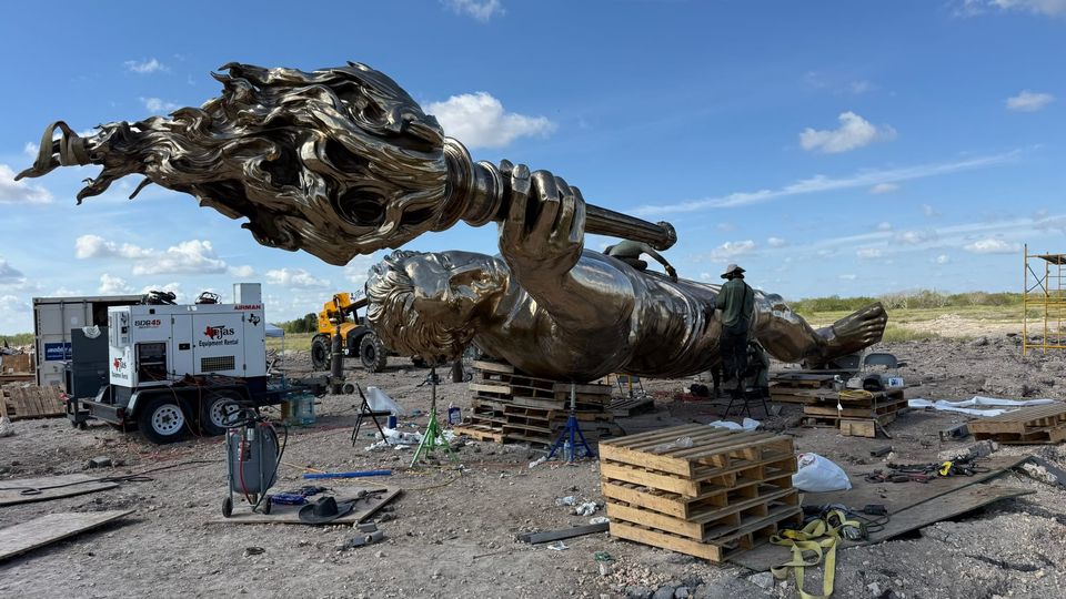 A giant bronze Prometheus statue lies on pallets at an outdoor worksite as workers assemble it