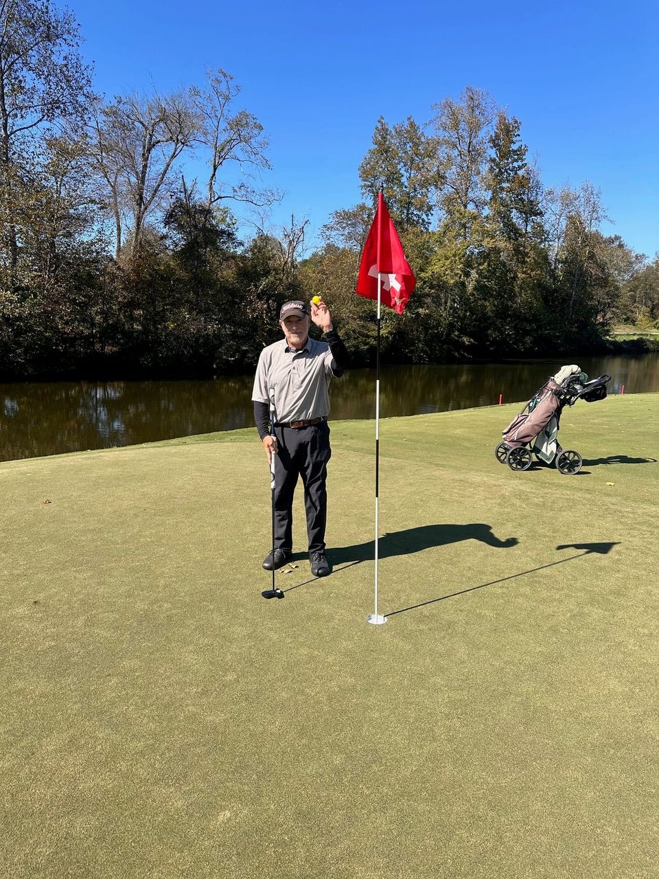 An older man holds up a golf ball beside a red flag on a sunny green, with a golf cart and pond behind him