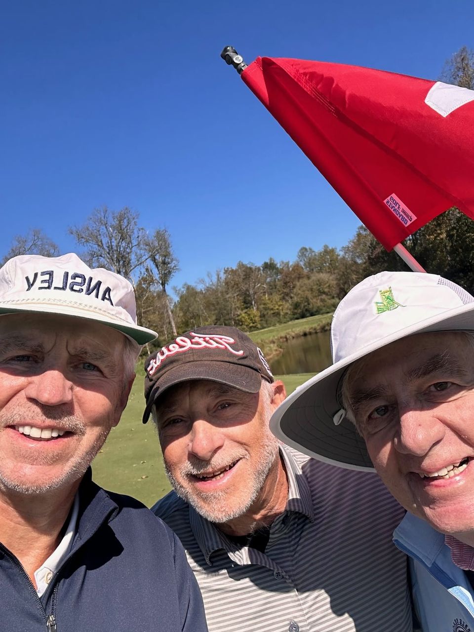 Three older men smile for a selfie on a golf course beneath a red flag