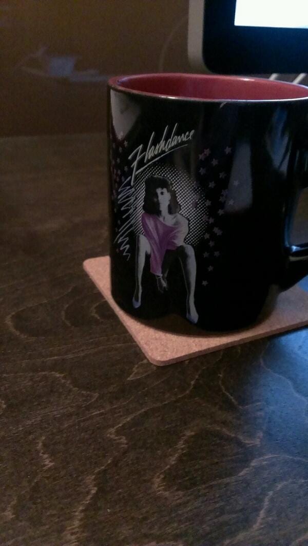 Black Flashdance mug with an illustration of a dancer in a pink off-shoulder top on a cork coaster