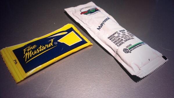 Yellow and navy Fine Mustard packet in retro script beside a white mayonnaise packet on a steel counter