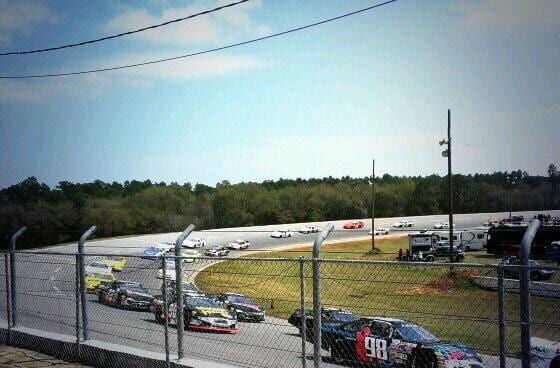 Stock cars racing around a banked short track seen through a chain-link fence