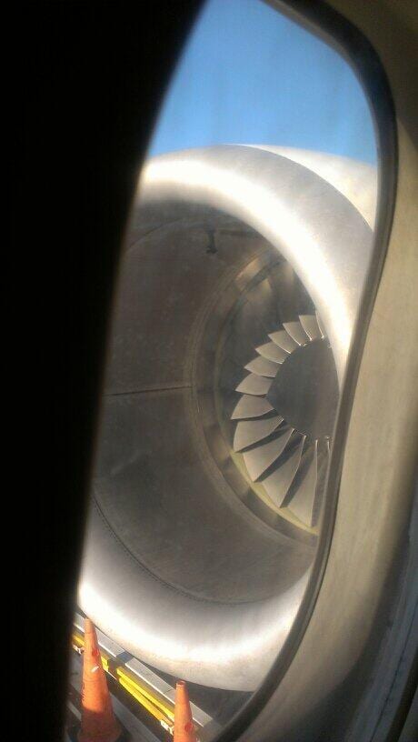 Jet engine intake with visible fan blades seen through an airplane window, orange cones below