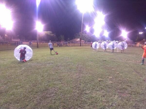 Players in inflatable bubble suits on a floodlit grass field at night as one kicks a ball