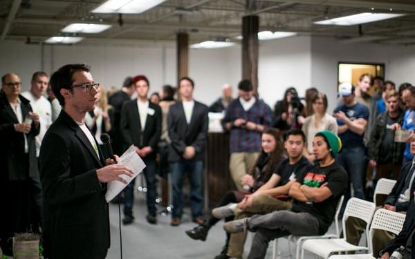 Man in glasses and a black blazer speaking into a microphone to a crowd in a loft event space