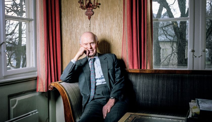 Elderly bald man in a gray suit seated on a sofa between red curtains, resting his chin on his hand