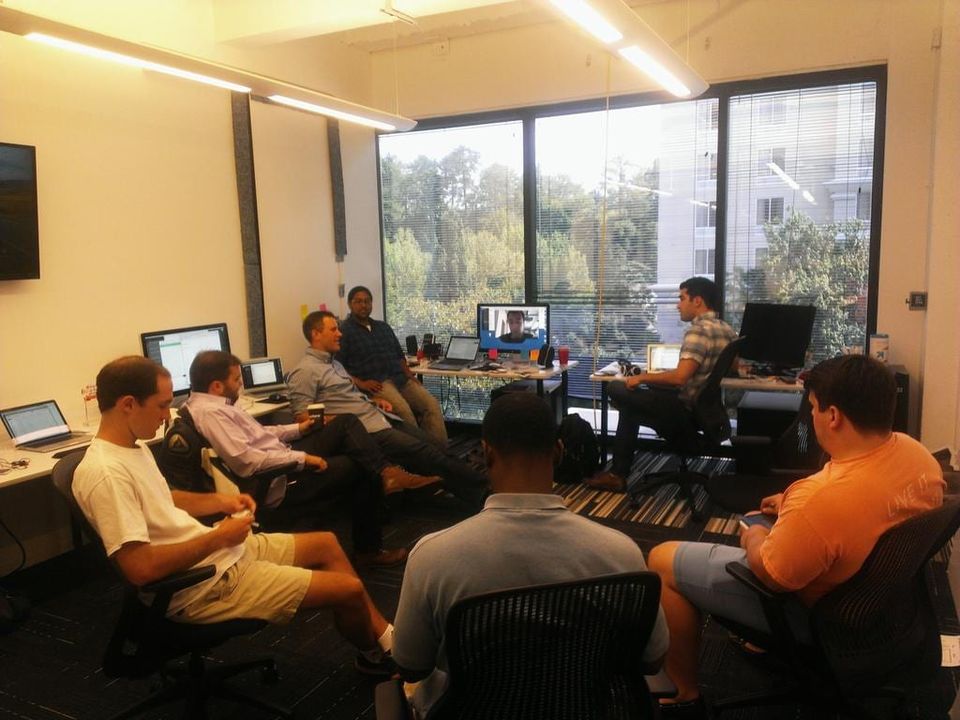 Group of men in office chairs circled for a meeting with a remote teammate on a screen by the windows