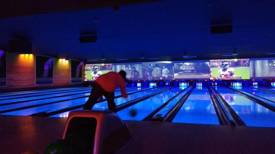 Bowler in an orange shirt releasing a ball down a blacklit lane beneath wall-wide screens showing football and a movie