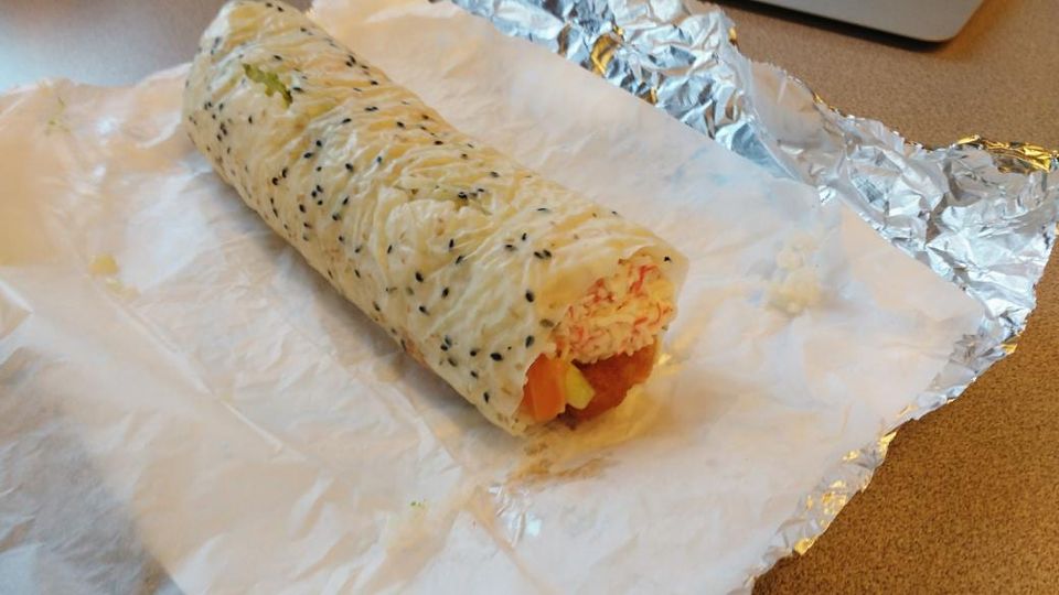 Sushi burrito dotted with black sesame seeds, cut open to show crab salad and vegetables, on paper and foil