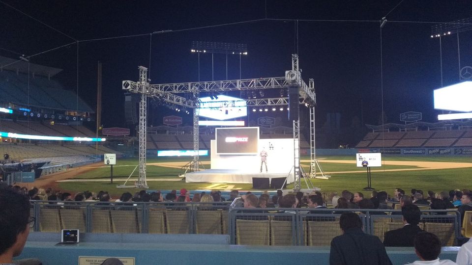 Presenter on a lit stage at home plate in Dodger Stadium with a SidePrize slide on screen and a seated crowd