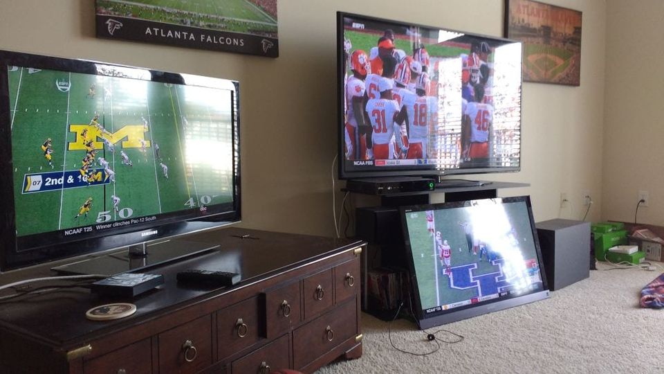 Three TVs in a living room each showing a different college football game beneath an Atlanta Falcons poster