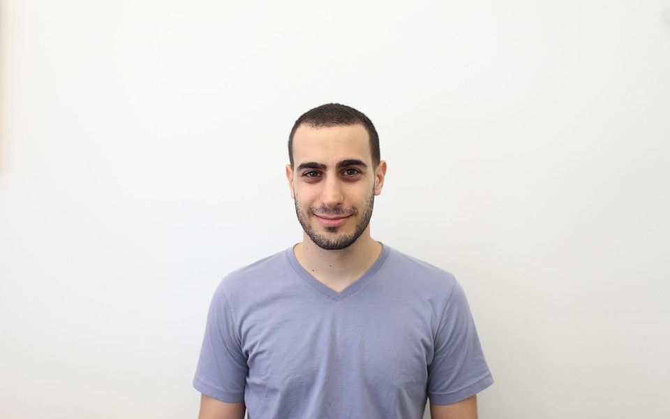 Man with a short beard in a lavender V-neck t-shirt smiling against a white wall