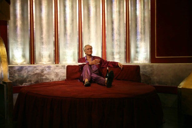 Wax figure of an older man in purple silk pajamas lounging on a round red bed with a bottle