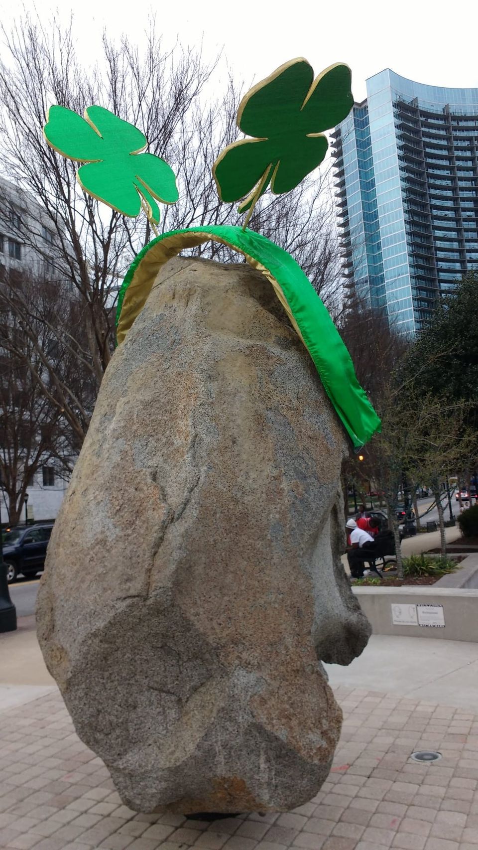 Large granite rock sculpture wearing a giant green shamrock headband on a plaza near high-rise towers