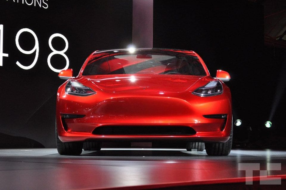 Front view of a red Tesla Model 3 with a smooth grille-less nose on a reveal stage