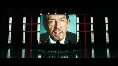 Film still of a man's face on a giant screen shouting over a dark stage