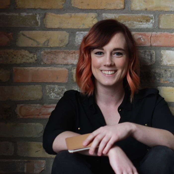 Smiling woman with wavy red hair in a black shirt sitting against a brick wall holding a phone