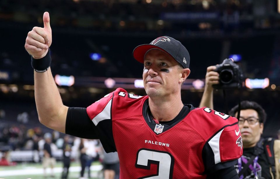 Falcons quarterback in a red number 2 jersey and cap giving a thumbs-up on the field as photographers follow