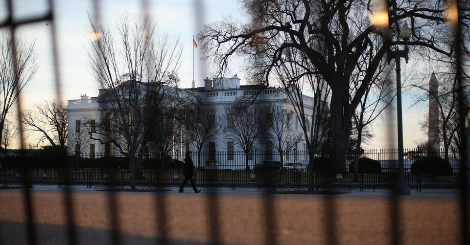 The White House at dusk seen through a blurred fence with a lone person walking past