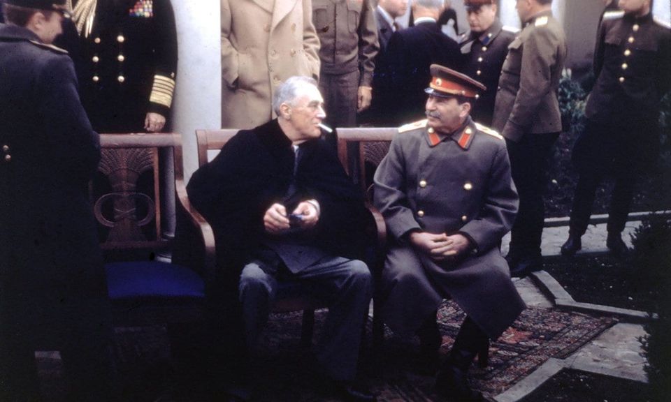 Color photo of Franklin Roosevelt in a dark cape seated beside Joseph Stalin in uniform, surrounded by officers
