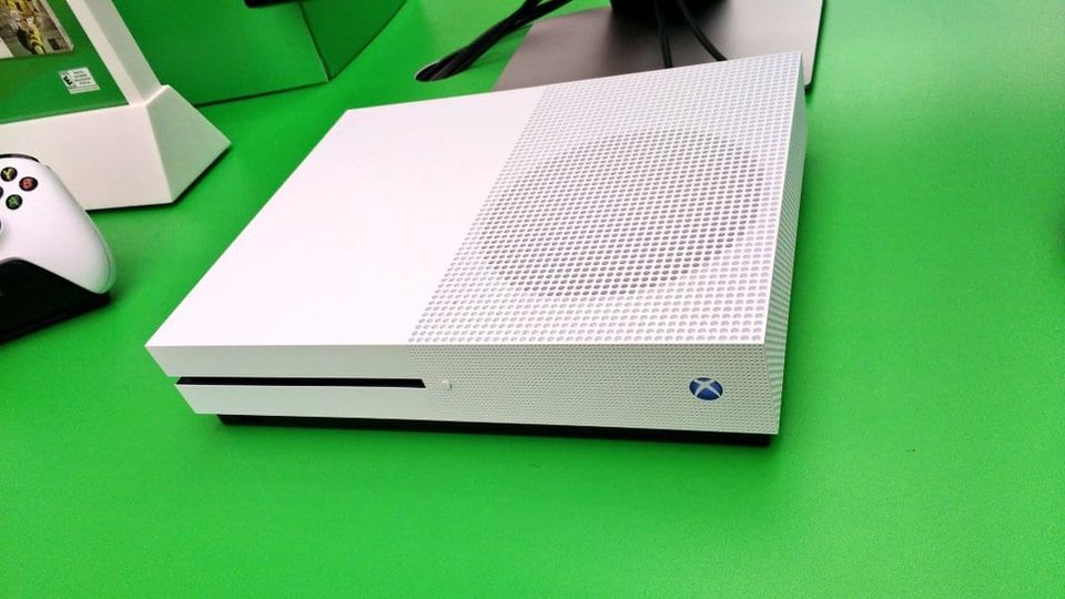 White Xbox One S console with a perforated top on a green display table beside a controller