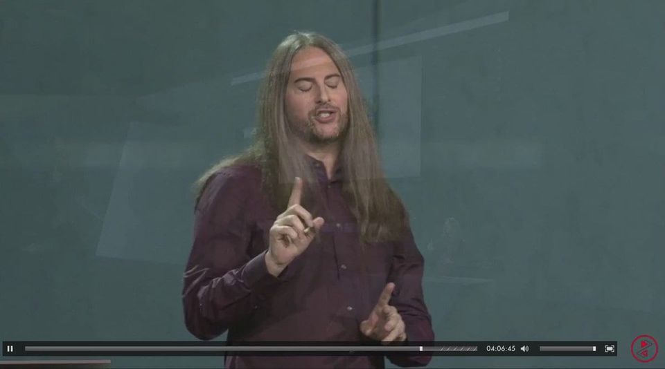 Long-haired presenter in a maroon shirt raising a finger mid-speech in a paused video at 04:06:45