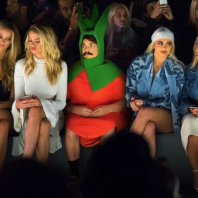 Mustached man in a carrot costume seated in a fashion show front row between glamorous women