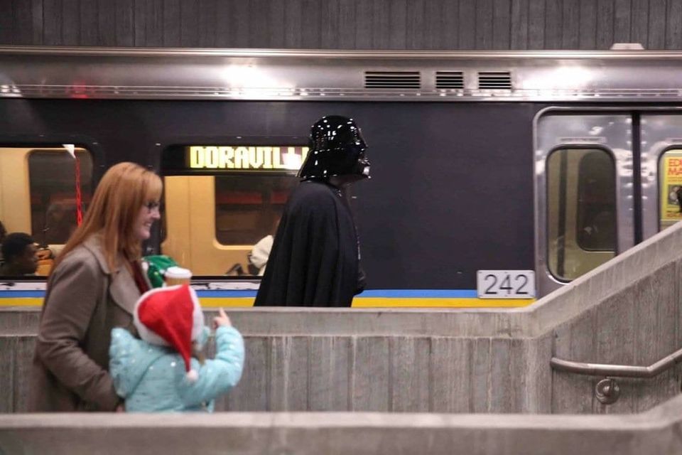 Person in a Darth Vader costume on a MARTA platform beside a Doraville train as a woman and child in a Santa hat pass