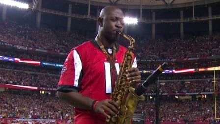 Man in a red Falcons jersey playing saxophone on the field of a packed stadium
