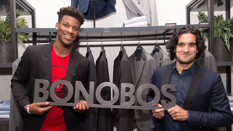 Two smiling men in blazers holding a large Bonobos sign in front of a rack of suits