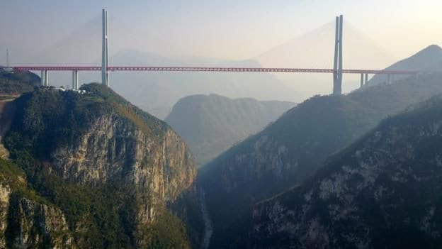 Red cable-stayed bridge spanning a deep misty gorge between two tall towers in the mountains