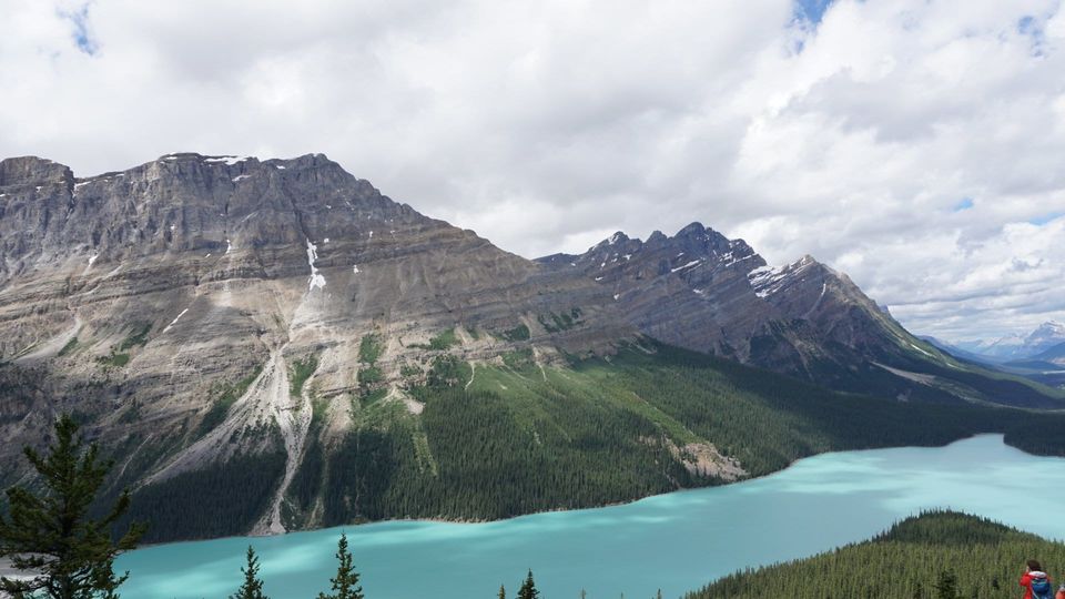 Turquoise glacial lake below rugged gray mountains and dense evergreen forest under a cloudy sky