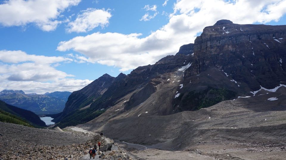 Hikers climbing a rocky ridge trail below dark cliffs with Lake Louise far below in the valley