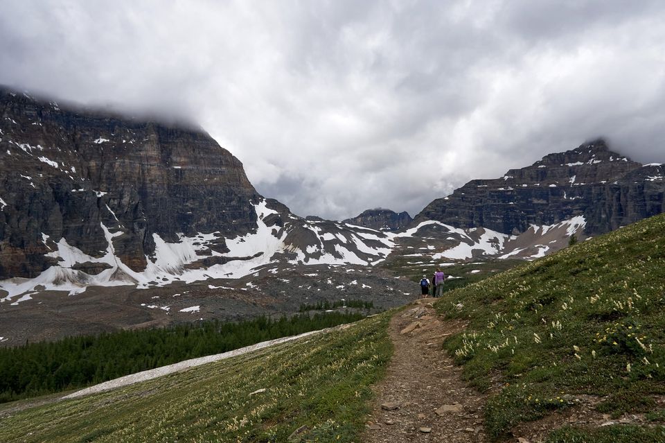 Hikers on a dirt trail through an alpine meadow toward cloud-shrouded cliffs and snowfields